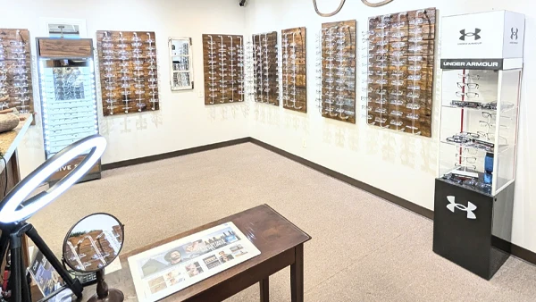 An optical store interior with multiple display walls showcasing a wide selection of eyeglass frames. A branded Under Armour eyewear stand is positioned on the right, and a table with marketing materials is in the foreground.